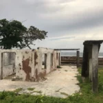 Weathered ruins amidst lush greenery and a cloudy sky, with a view of the ocean in the distance at French Cay, Roatán, Honduras.