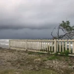 Stormy skies loom over the ocean near a white picket fence in Oak Ridge, Roatán Island, Honduras, with waves crashing nearby.