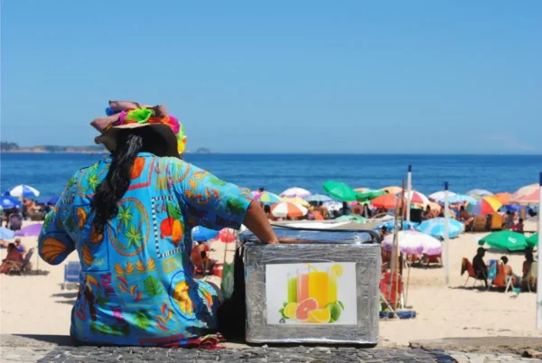 Pessoa com camisa e chapéu coloridos sentada ao lado de um isopor de sucos, de frente para o mar e guarda-sóis