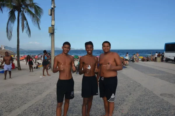 Três jovens brasileiros sem camisa sorrindo e fazendo sinal de positivo perto do mar, em uma passarela lotada