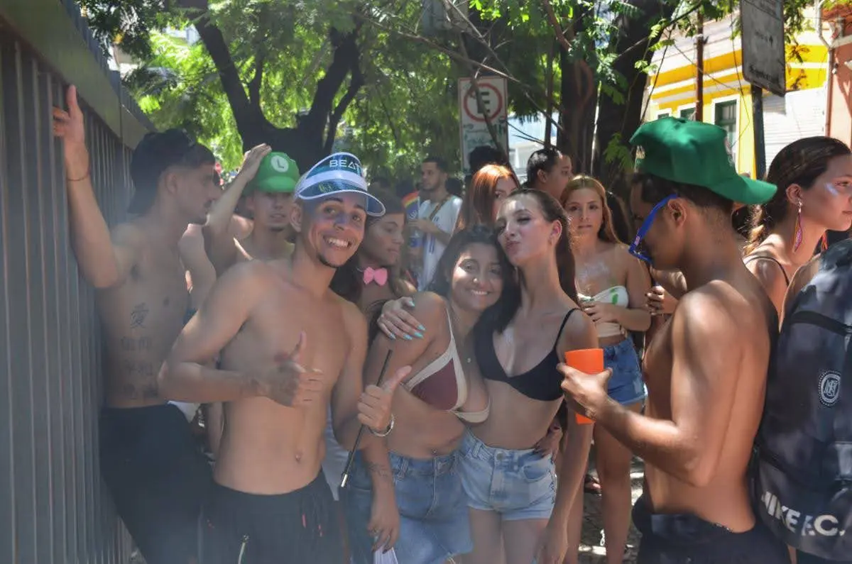 Group of young people posing and smiling during a Rio de Janeiro street party for Carnival, with shirtless men and women in crop tops and denim shorts gathered under leafy trees in bright daylight