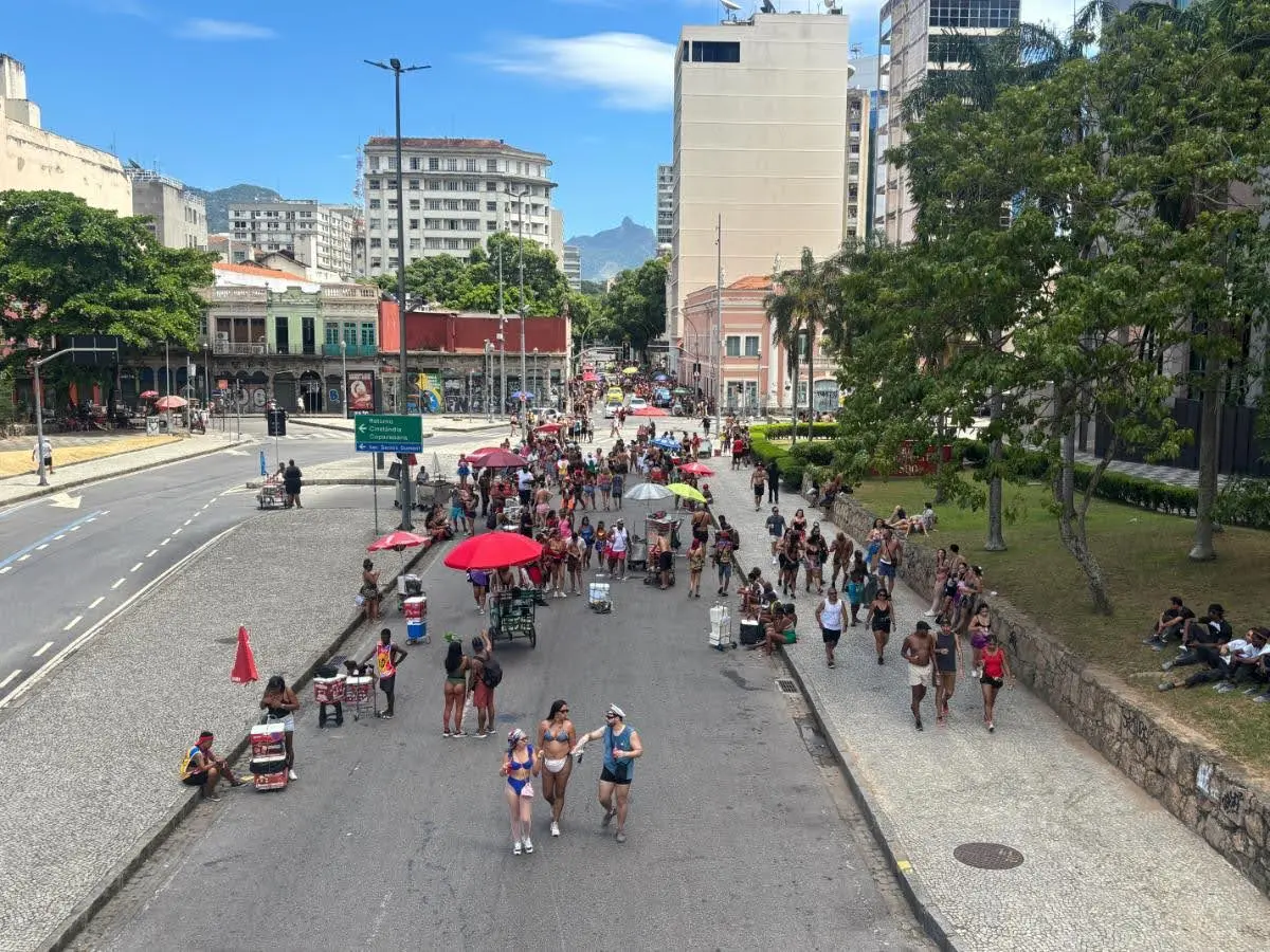 Mass of people dancing and walking in a downtown Rio street during a daytime Carnival bloco, surrounded by buildings and bright decorations