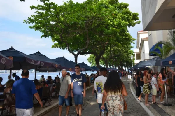 Umbrella-covered cafe tables and pedestrians walking along a tree-lined promenade near Fort Copacabana, with beach views and local restaurants creating a lively daytime atmosphere.