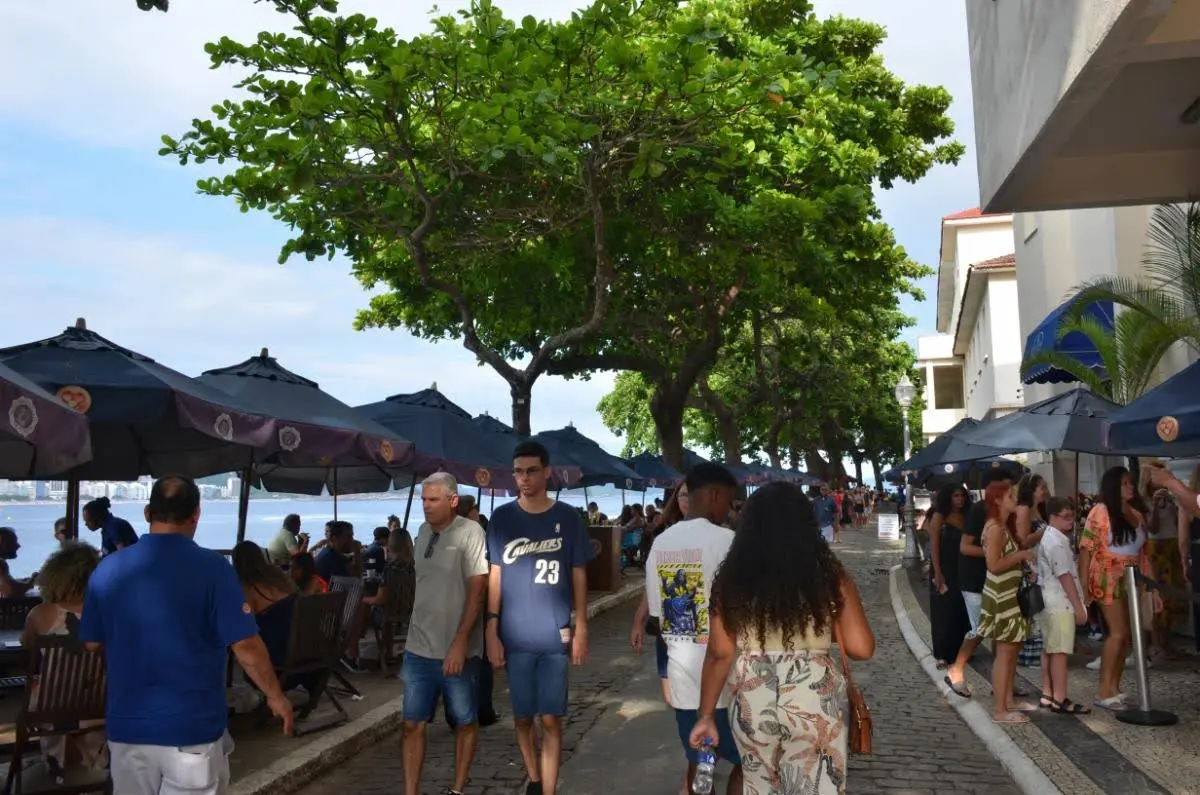 Umbrella-covered cafe tables and pedestrians walking along a tree-lined promenade near Fort Copacabana, with beach views and local restaurants creating a lively daytime atmosphere.