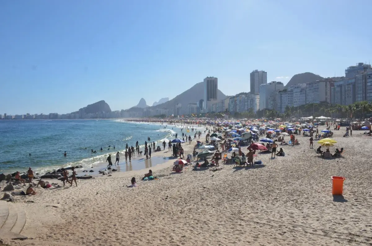 Wide-angle shot of Copacabana Beach in Rio de Janeiro with beachgoers, high-rise buildings, and iconic sidewalks stretching along the bustling shoreline