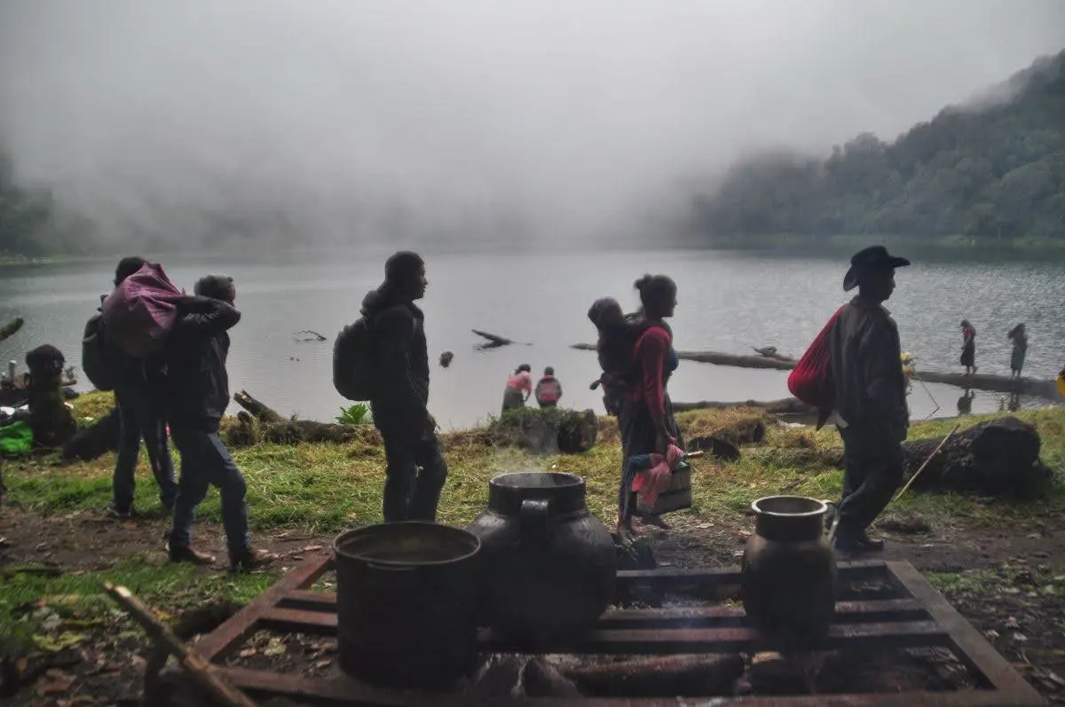 Pilgrims walking in front of platform with steaming kettles on shore of laguna chicabal