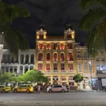 Historic colonial building illuminated at night in the Lapa neighborhood of Rio de Janeiro, framed by tall palm trees and vibrant street life, capturing the district’s architectural charm and festive energy.