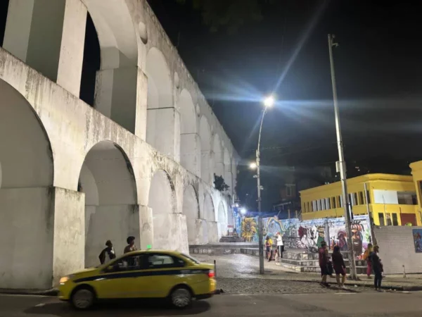 The Arcos da Lapa aqueduct in Rio de Janeiro at night, dramatically lit and supporting a yellow tram overhead, with pedestrians and cars passing beneath—symbolizing the district’s cultural and historic center.