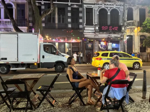 Locals and tourists gather at outdoor tables along a brightly lit street in Lapa, Rio de Janeiro, lined with bars, music, and vibrant nightlife activity, setting the stage for an energetic night out.