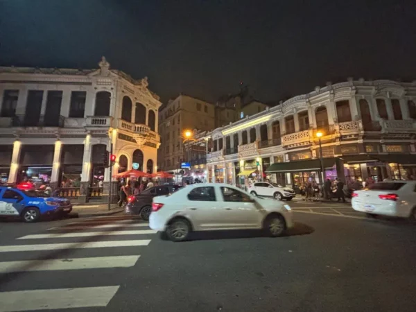 Classic colonial façades and neon-lit shops line a busy corner in Lapa, Rio de Janeiro, as cars cruise by and people gather outside bars—showing the lively blend of history and nightlife that defines the district.