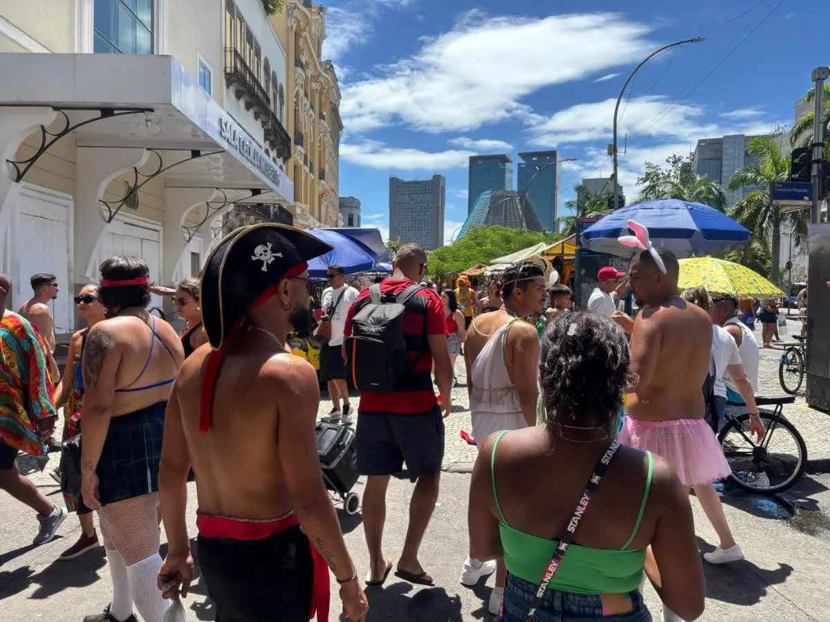 Carnival crowd in Rio de Janeiro with people dressed in playful costumes including pirate hats, tutus, bunny ears, and schoolgirl skirts, gathered under bright sun and umbrellas on a downtown street with modern skyscrapers in the background