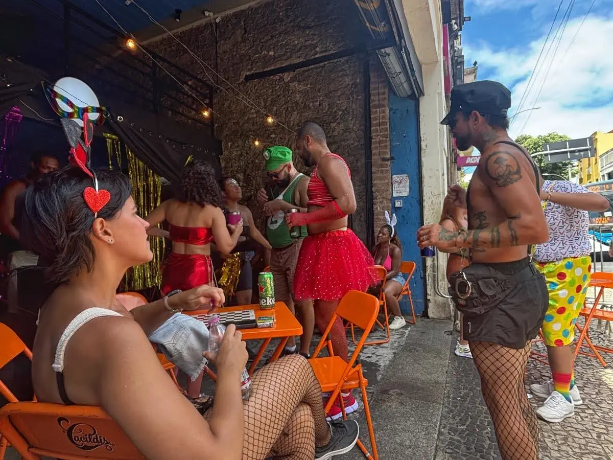 Carnival-goers relaxing outside a bar in Rio de Janeiro, wearing fishnet tights, colorful skirts, and festive accessories while chatting around bright orange chairs and tables on a cobblestone alley during Carnival celebrations