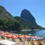 Scenic view of Sugarloaf Mountain towering above the shoreline at Praia Vermelha in Rio de Janeiro, with a sunny sky, clear water, and vibrant beach umbrellas in the foreground