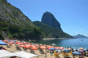 Scenic view of Sugarloaf Mountain towering above the shoreline at Praia Vermelha in Rio de Janeiro, with a sunny sky, clear water, and vibrant beach umbrellas in the foreground