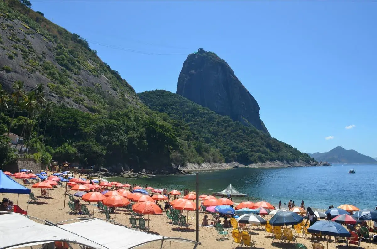 Scenic view of Sugarloaf Mountain towering above the shoreline at Praia Vermelha in Rio de Janeiro, with a sunny sky, clear water, and vibrant beach umbrellas in the foreground