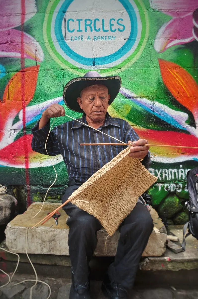 guatemalan man with cowboy hat Knitting in the street sat on ledge in front of mural in san marcos la laguna