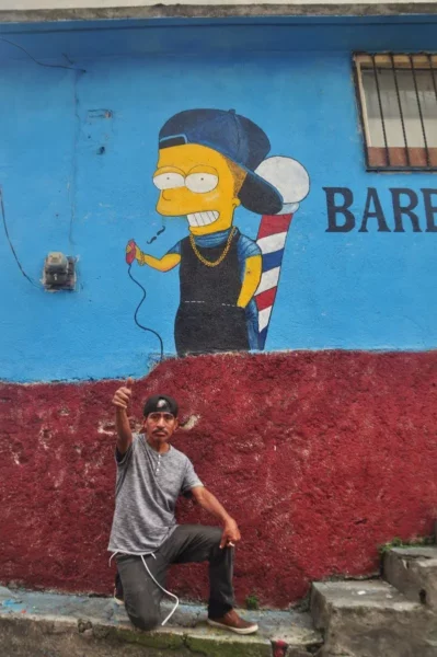 Street mural of a Bart-Simpson style character on a blue wall, with a man posing on the steps in San Pedro La Laguna