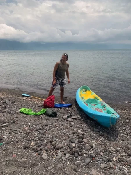 Man with a blue kayak on the rocky shore of Lake Atitlán, getting ready to paddle under cloudy skies