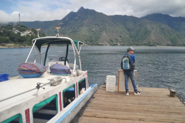 Small passenger boat at a wooden dock on Lake Atitlán, with a traveler waiting and volcanoes in the background.