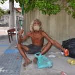 Homeless man in Cracolândia sitting shirtless on the sidewalk beside black trash bags and a shopping cart, symbolizing the area's extreme poverty and street life