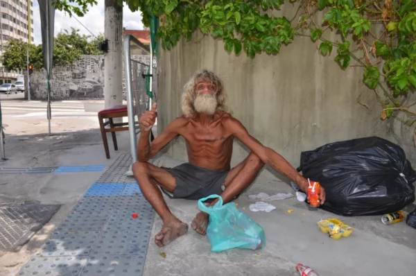 Homeless man in Cracolândia sitting shirtless on the sidewalk beside black trash bags and a shopping cart, symbolizing the area's extreme poverty and street life