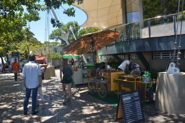 Área de lanchonetes ao ar livre no topo do Morro da Urca, com mesas, guarda-sóis e barracas oferecendo comidas e bebidas