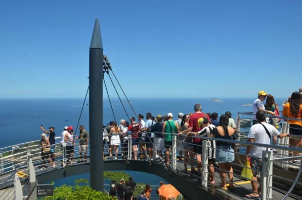 Turistas em uma passarela curva no topo do Pão de Açúcar, com vista panorâmica do litoral e céu do Rio de Janeiro