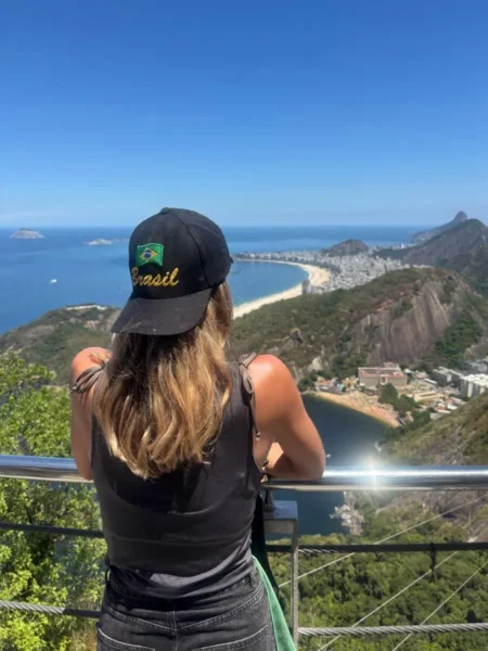 Mulher com boné da bandeira do Brasil observa a vasta costa do Rio de Janeiro do alto do Pão de Açúcar, com céu azul e vista do mar em direção a Copacabana