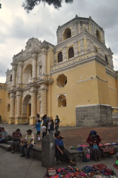 Yellow baroque façade of Iglesia La Merced in Antigua Guatemala, with street vendors sitting in front of the church