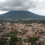 Aerial view of Antigua Guatemala with the colorful colonial grid and Volcán de Agua rising under a cloudy sky from Cerro de la Cruz
