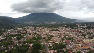 Aerial view of Antigua Guatemala with the colorful colonial grid and Volcán de Agua rising under a cloudy sky from Cerro de la Cruz