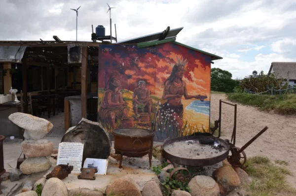 A vibrant mural of indigenous figures adds color and meaning to this rustic beachside eatery, surrounded by weathered cooking pots and stones that hint at stories of the land and fire.