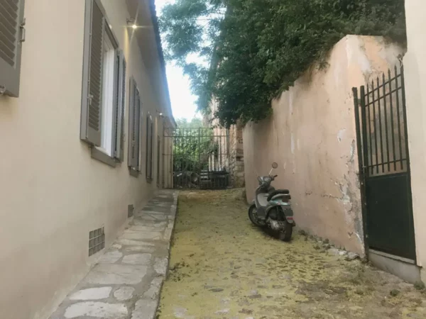 Narrow alleyway lined with warm-toned buildings and overgrown foliage, leading to a gated entrance in Chalkis, Euboea, Greece.