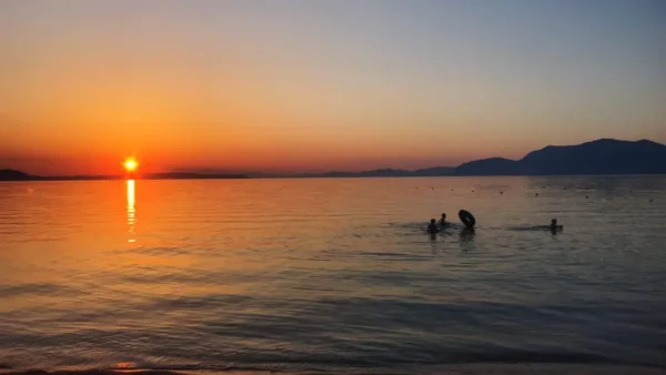 Vibrant sunset casts golden hues over calm waters, with silhouettes of people swimming near the shore in Chalkis, Euboea, Greece.