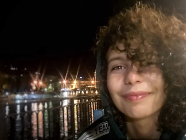 Chalkis sparkles at night, reflecting colorful lights on the water as a smiling visitor enjoys the view.