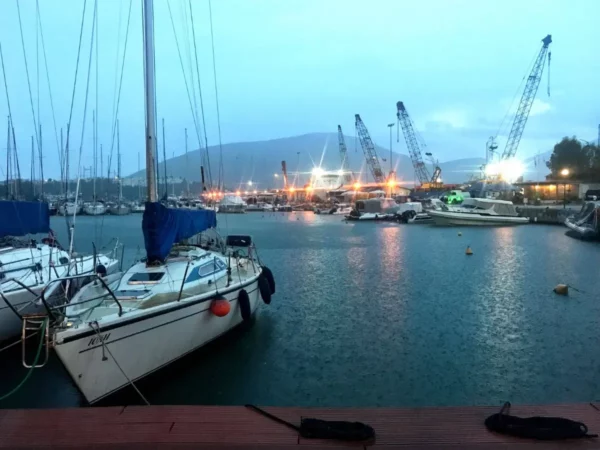 Sailboats with blue sails docked at a marina, surrounded by cranes and a misty dusk sky in Chalkis, Euboea, Greece.