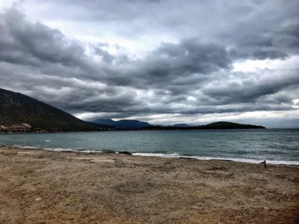 Stormy clouds hang over the sandy beach of Chalkis, Euboea, with gentle waves lapping at the shore and distant mountains visible.