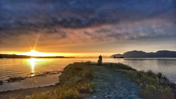Golden sunrise over the water with dark clouds, casting reflections near Chalkis, Euboea, Greece. Coastal path lined with plants.