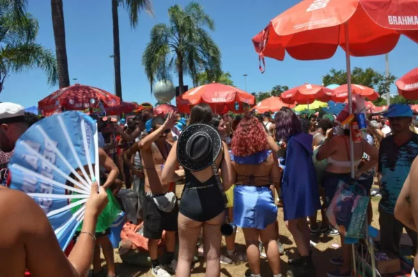 Crowd gathered at Rio de Janeiro Bloco do Sargento Pimenta Carnival street party, with people in creative costumes dancing under red umbrellas and palm trees, some fanning themselves in the midday heat