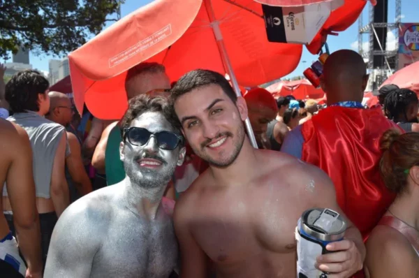 Two men posing during Rio Carnival, one covered in silver body paint and wearing sunglasses, the other shirtless and smiling, surrounded by red umbrellas and fellow revelers at a busy street party