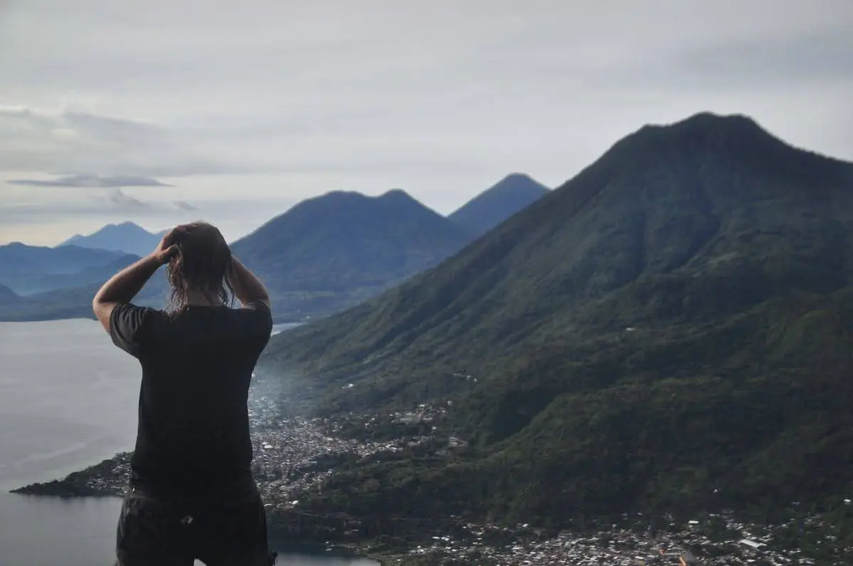 Person standing at a viewpoint above Lake Atitlán, looking out over San Juan La Laguna with steep green hills and volcanoes rising along the shoreline under a cloudy sky.