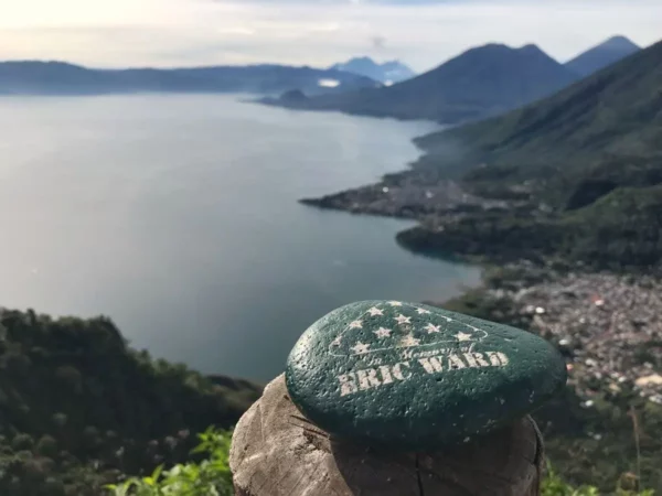 Green painted rock with "Eric Ward" sits atop a scenic viewpoint overlooking Lake Atitlán, surrounded by lush hills and distant mountains.