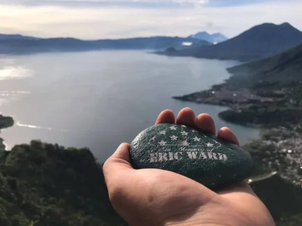 Hand holding a engraved stone with "Eric Ward" overlooking Lake Atitlán, surrounded by lush hills and distant mountains in Guatemala.