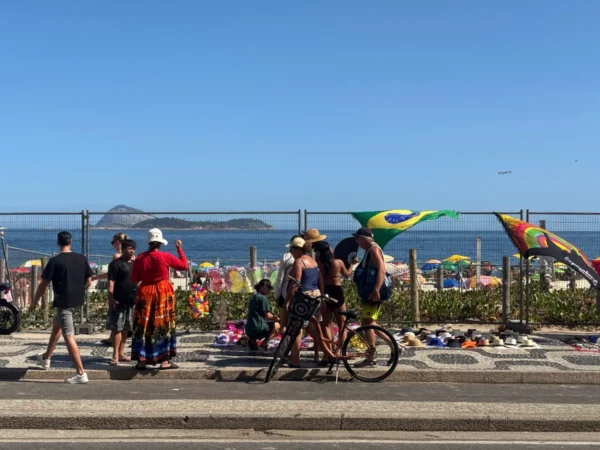 Um grupo de pessoas e ambulantes reunidos perto da grade da Praia de Ipanema, com bandeiras do Brasil e vista para o mar ao fundo