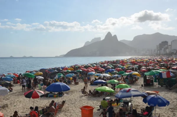 Praia de Ipanema lotada com guarda-sóis coloridos, banhistas e o contorno das montanhas Dois Irmãos ao fundo
