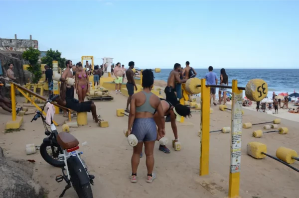 Pessoas malhando em uma academia ao ar livre próxima à Praia de Ipanema, com halteres de concreto e bancos improvisados à beira-mar
