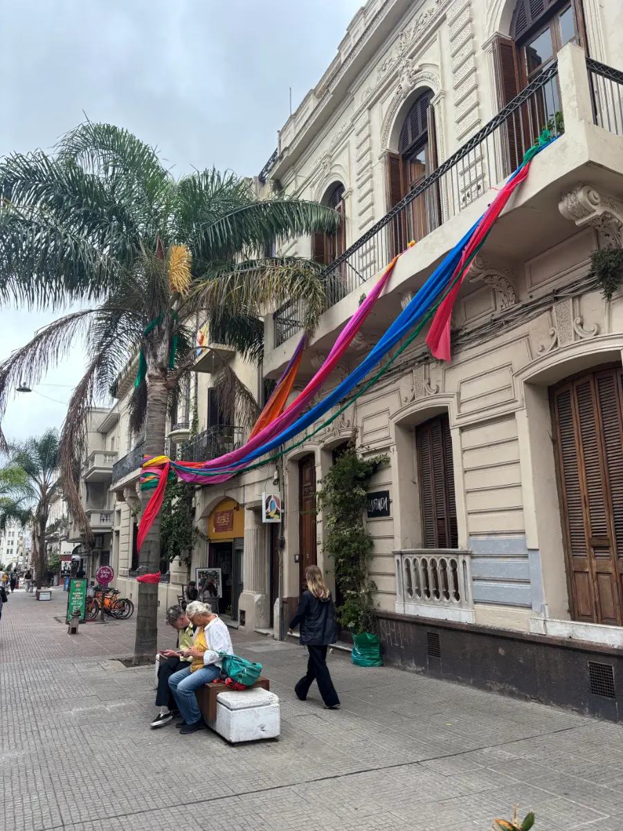 Colorful fabric streamers drape between palm trees and colonial façades on a lively street in montevideo old city, as people relax on a bench below.