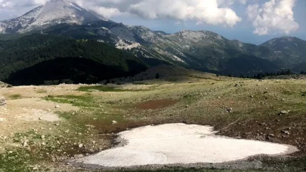 Snowy patches contrast with the green landscape and rocky terrain of Mount Xerovouni in Euboea, Greece, under a cloudy sky.