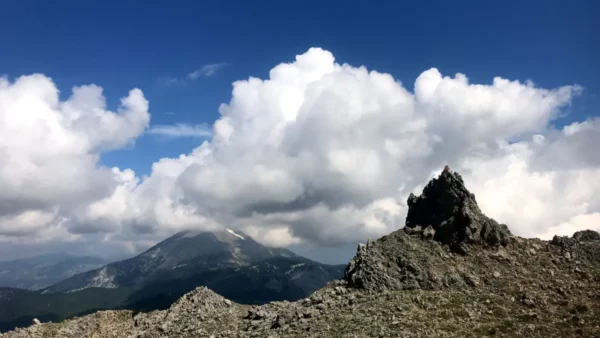 Lush green slopes and rugged rock formations under a bright blue sky with fluffy clouds at Mount Xerovouni, Euboea, Greece.
