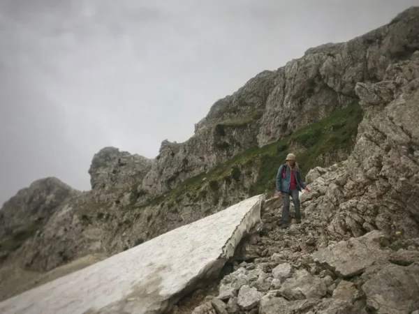 A hiker navigates rocky terrain on Mount Xerovouni, Euboea, Greece, with rugged cliffs and a cloudy sky in the background.